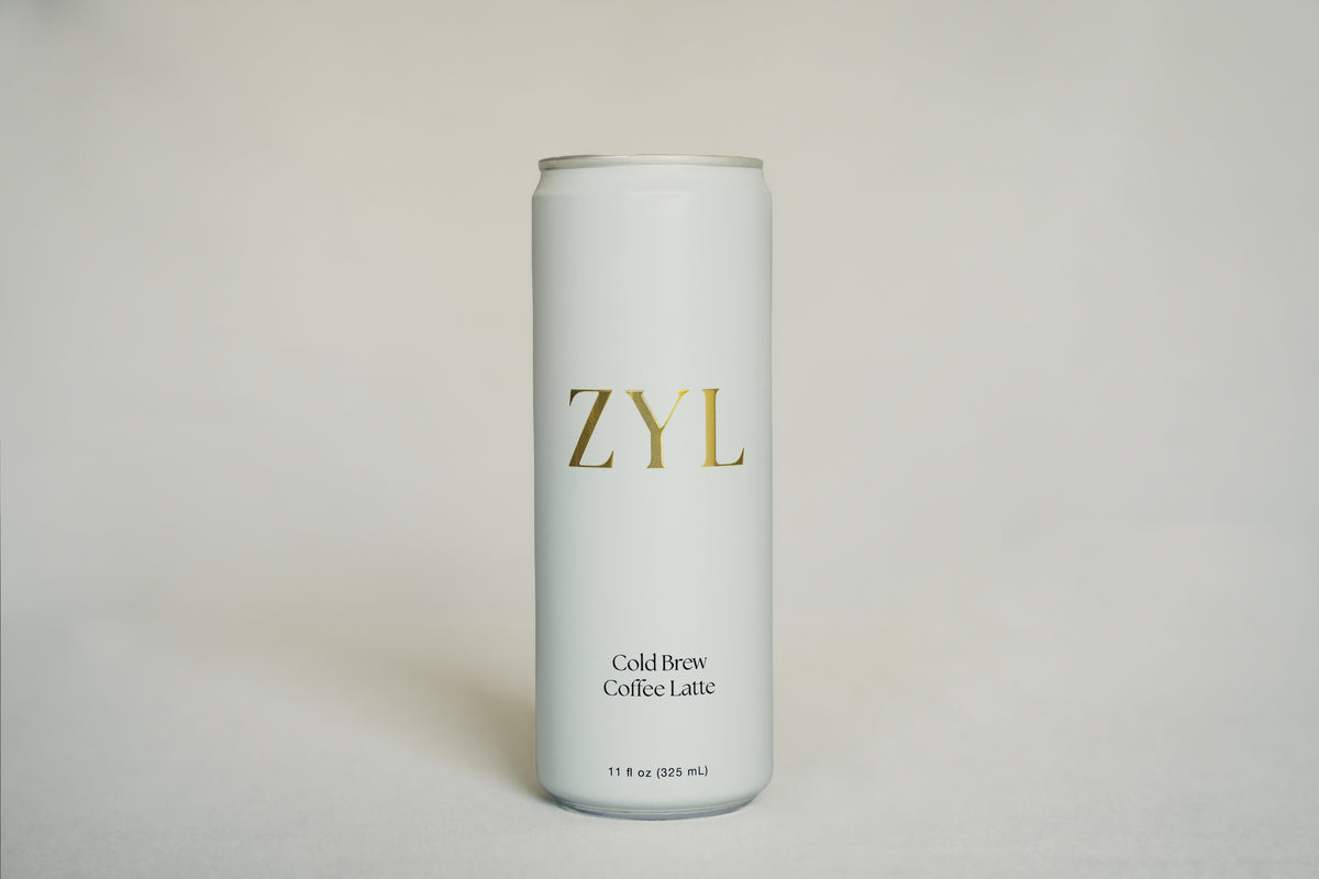 A tall, slim aluminum can of ZYL Cold Brew Coffee Latte, featuring a minimalist white design with gold and black lettering, set against a neutral light cream background.