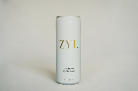 A tall, slim aluminum can of ZYL Cold Brew Coffee Latte, featuring a minimalist white design with gold and black lettering, set against a neutral light cream background.